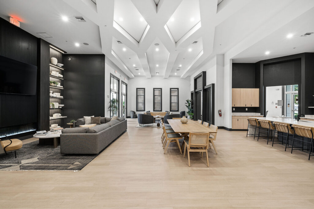 Spacious, modern lounge area with gray sofas, a long wooden dining table, bar seating, open shelves, large windows, and high ceilings with skylights. The space features minimalist decor and neutral tones.