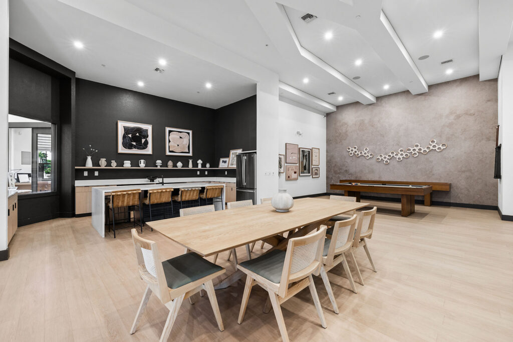 Modern communal room with a long wooden dining table and chairs, a kitchen area with barstools, wall art, decorative shelves, and a pool table against a textured wall. The space is well-lit with high ceilings.