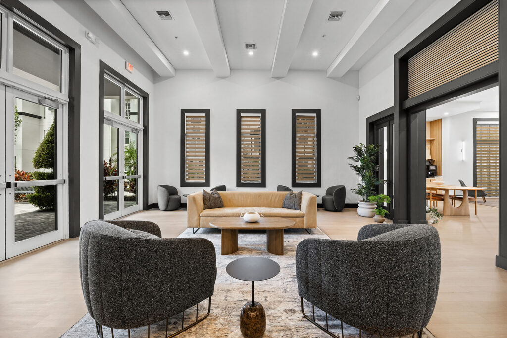 Modern, stylish lobby with two gray armchairs, a tan curved sofa, a wooden coffee table, and potted plants. Large windows and neutral decor create a bright, inviting atmosphere.