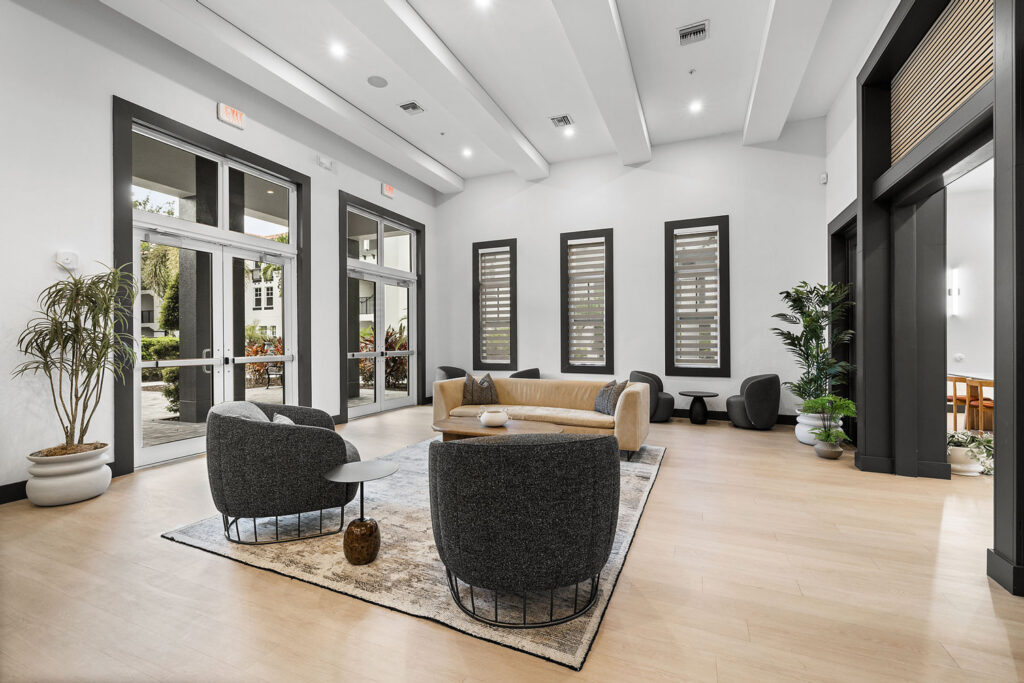 Modern lobby with light wood flooring, neutral tones, a beige sofa, two dark gray chairs, potted plants, and large windows letting in natural light. Contemporary decor and a cozy seating area on a patterned rug.