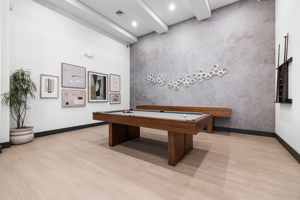Modern game room with a wooden pool table, abstract art on white and gray walls, a plant in the corner, a wall-mounted cue rack, and light wood flooring under bright recessed lighting.