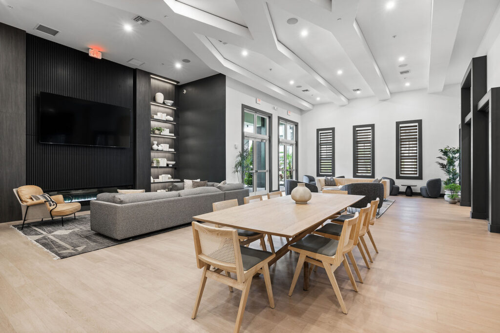 Modern, spacious lounge area with light wood flooring, gray sofas, a large wall-mounted TV, built-in shelves, a long wooden dining table with chairs, and large windows letting in natural light.