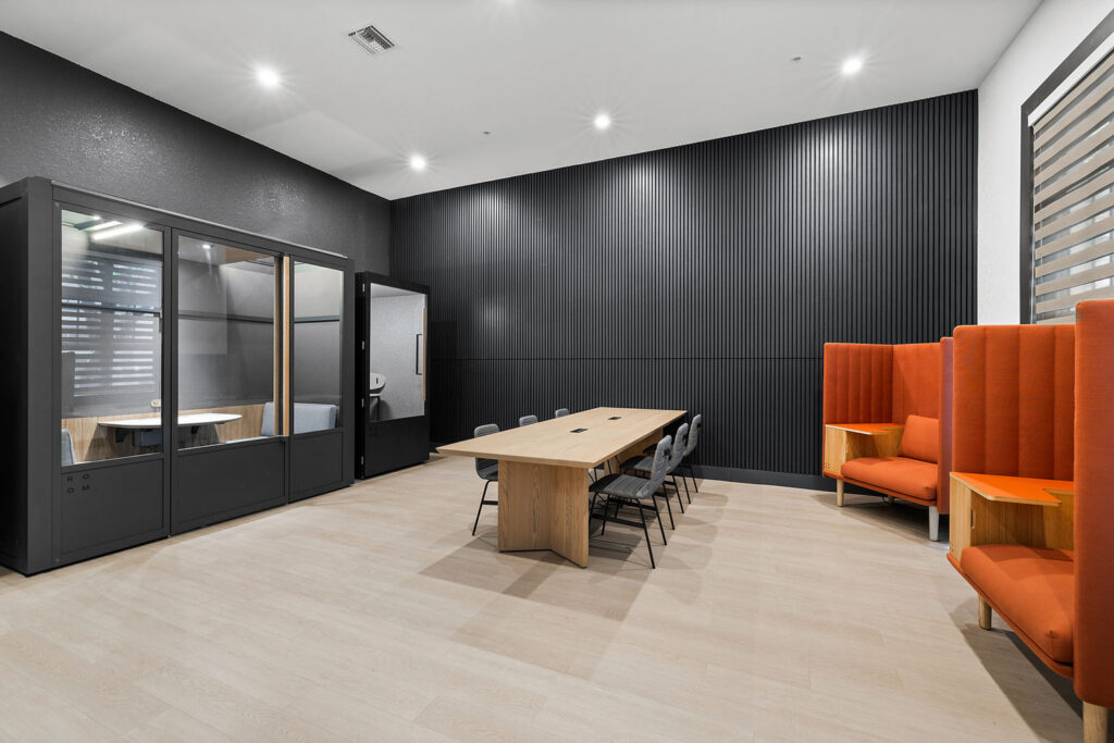 Modern conference room with a long wooden table, black chairs, two orange high-back sofas, black accent walls, and enclosed glass pods on one side. The room has light wood flooring and minimalistic decor.