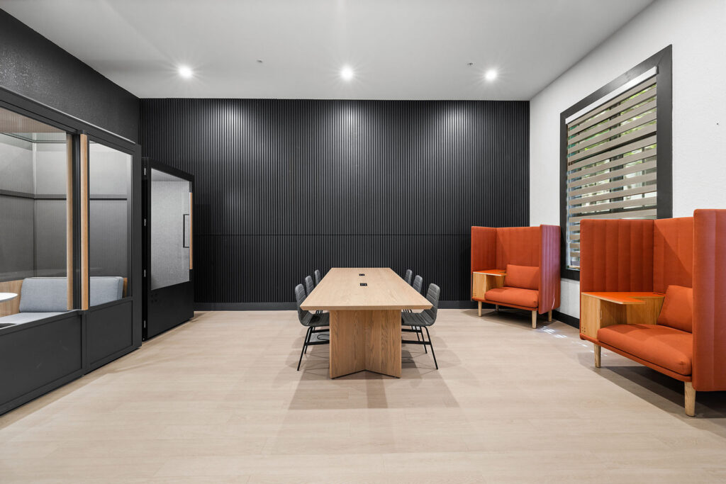 Modern conference room with a long wooden table surrounded by black chairs, two orange high-back chairs with wooden side tables, a glass meeting booth, black accent wall, and large window with blinds.
