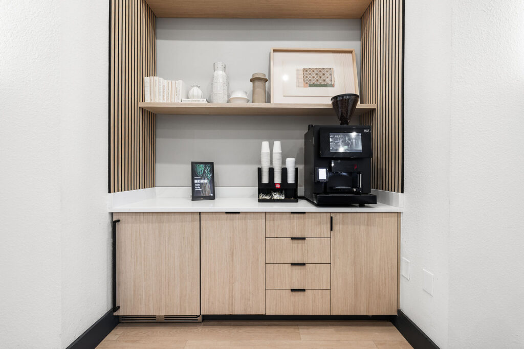 A modern coffee station with a black coffee machine, stacked cups, and stirrers on a white countertop with light wood cabinets below and decorative items on a shelf above.