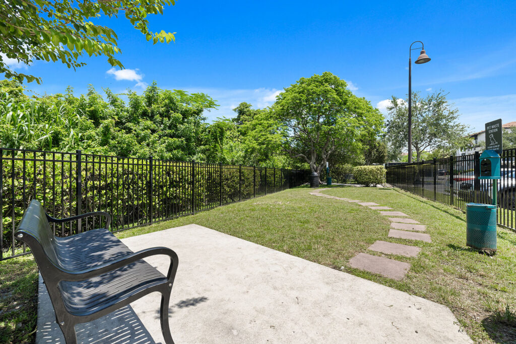 A small fenced-in park area with a black metal bench, a green waste bin, a dog waste station, a stone path, and lush trees under a bright blue sky.