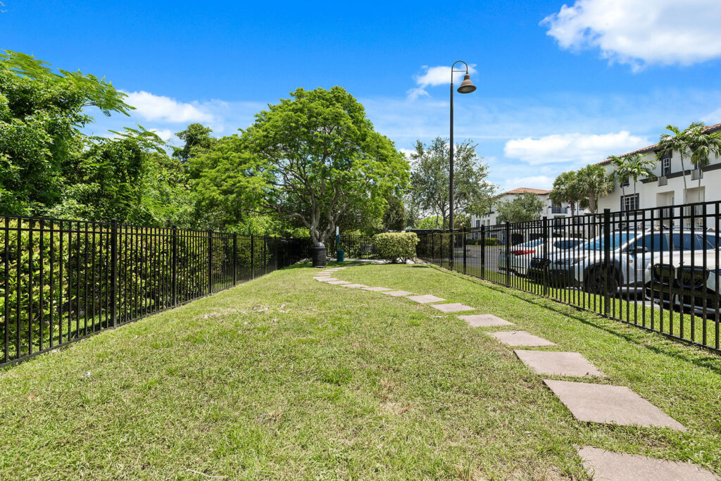 A grassy area with a winding stone path, bordered by a black metal fence on the left, trees and bushes in the background, and parked cars on the right under a bright blue sky with scattered clouds.