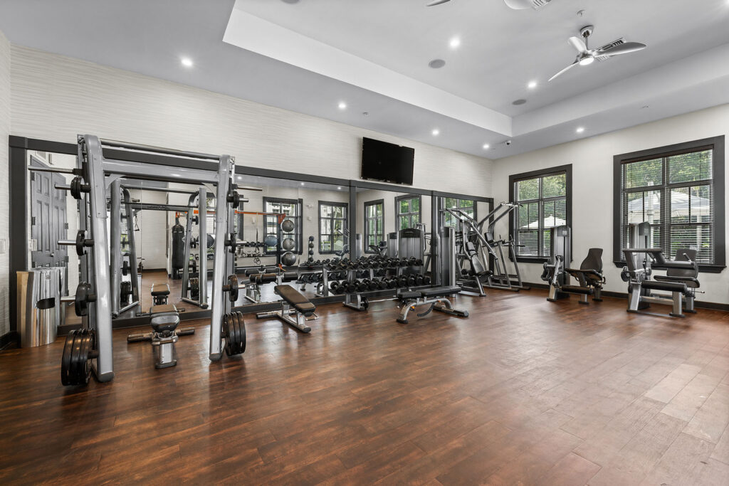 A modern gym with wood flooring, large mirrors, weightlifting machines, free weights, benches, treadmills, and exercise bikes. Windows let in natural light, and the space appears clean and well-lit.