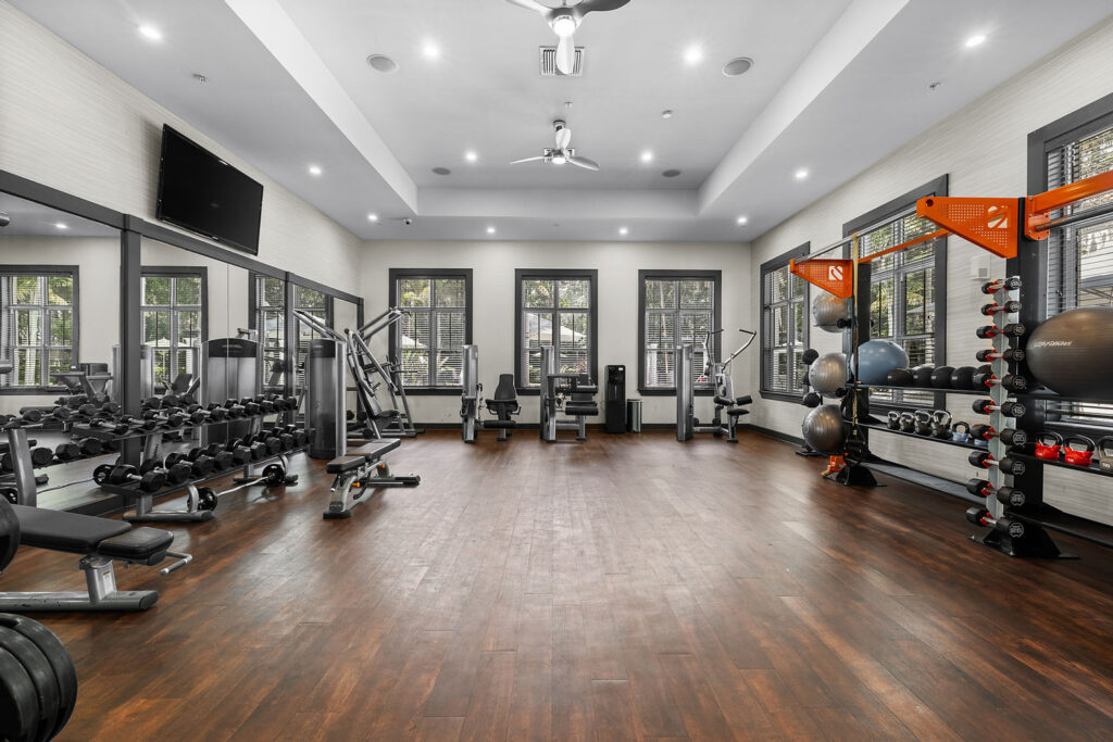 A spacious, well-lit gym with large windows, wood flooring, free weights, benches, weight machines, exercise balls, and wall-mounted racks, creating a modern and organized workout environment.