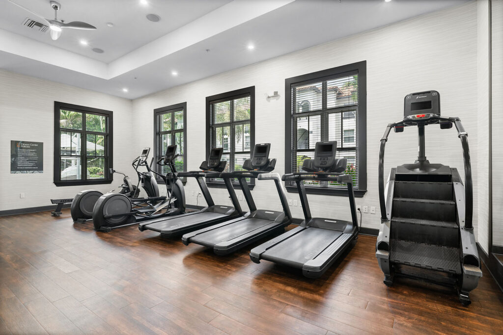 A modern gym with wooden floors, four cardio machines including two treadmills, an elliptical, and a stair climber, positioned next to large windows letting in natural light.
