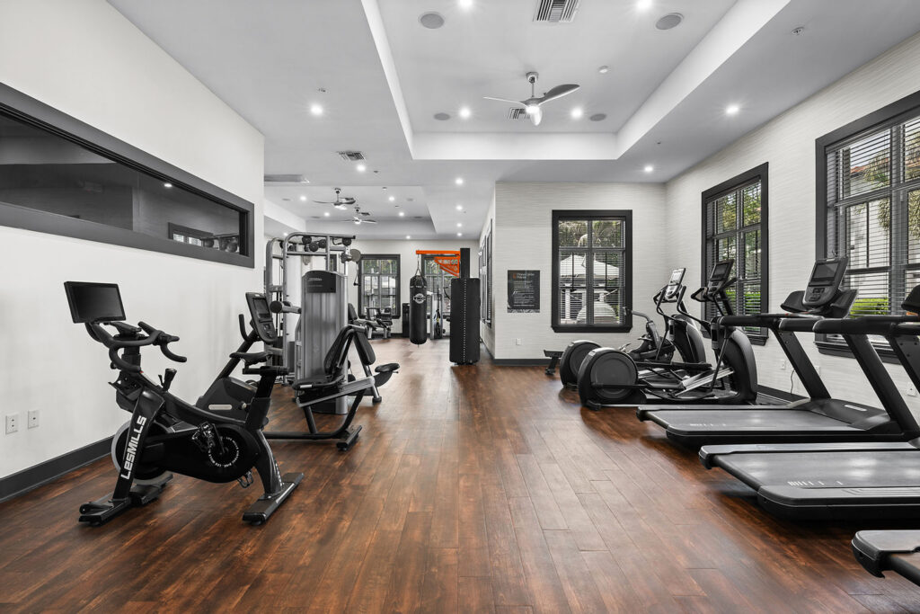 A modern gym with wooden floors, several treadmills, elliptical machines, stationary bikes, and strength training equipment, with large windows letting in natural light.