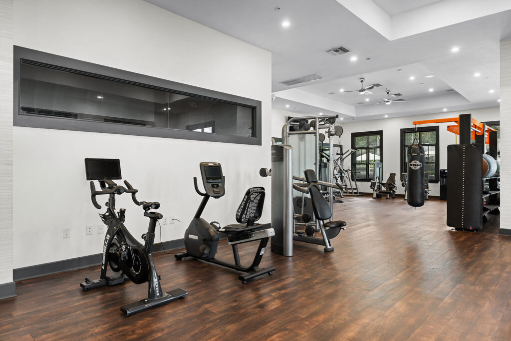 Modern gym with hardwood floors, featuring stationary bikes, a weight machine, treadmills, and various exercise equipment. Large windows let in natural light, and mirrors line the far wall.