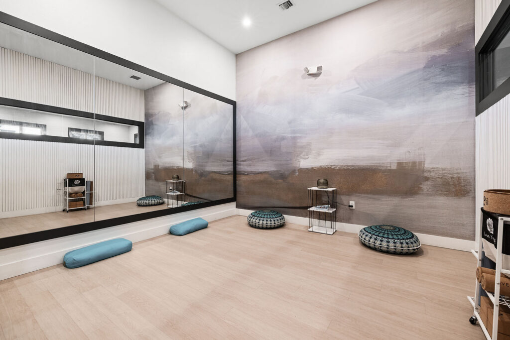 A modern yoga or meditation room with light wood floors, two blue cushions, two patterned floor pillows, a large wall mirror, soft abstract wall art, and small shelving units holding supplies.