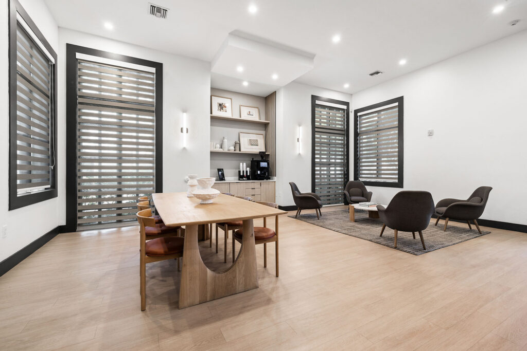 A modern lounge area with a wooden dining table and chairs, a coffee station, and a small sitting area with four armchairs around a rug. Large windows with horizontal blinds let in natural light.