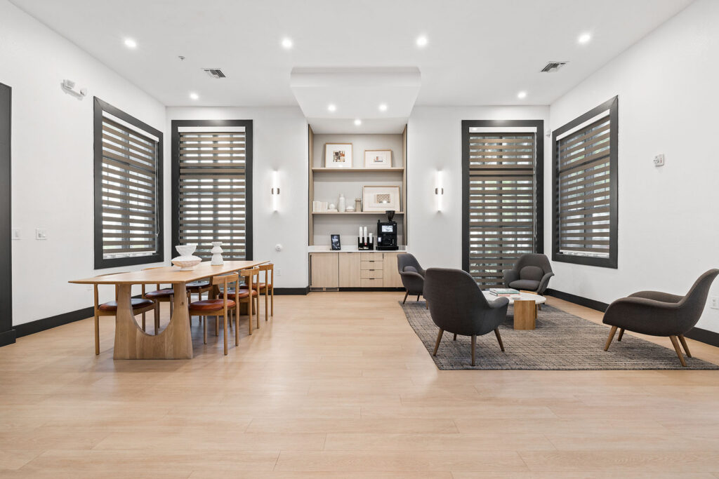 Modern lounge with light wood floors, a dining table with six chairs on the left, a cozy seating area with four gray armchairs and a small table on the right, and large windows with striped blinds throughout the room.