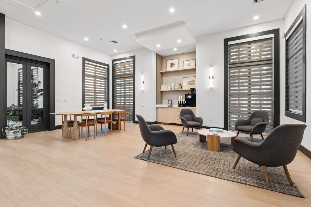 Modern lounge area with four dark armchairs around a small coffee table on a rug, a dining table with six chairs, wooden floors, large windows with striped blinds, and a built-in coffee station on the far wall.