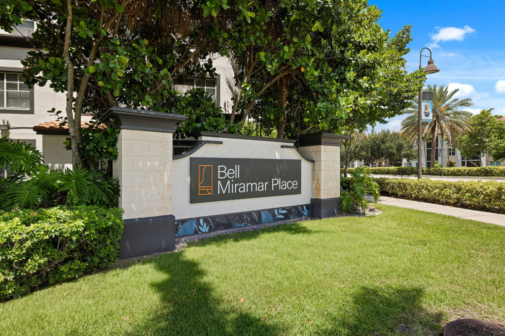 A large sign reading Bell Miramar Place stands on a well-manicured lawn, surrounded by lush greenery and trees, with a sidewalk and street lamp visible in the background.
