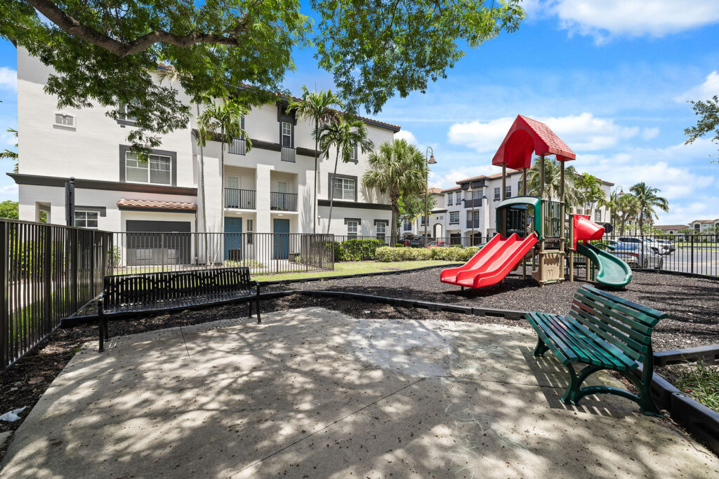 A fenced playground with red slides and green equipment sits next to two benches, with apartment buildings and palm trees in the background under a partly cloudy sky.
