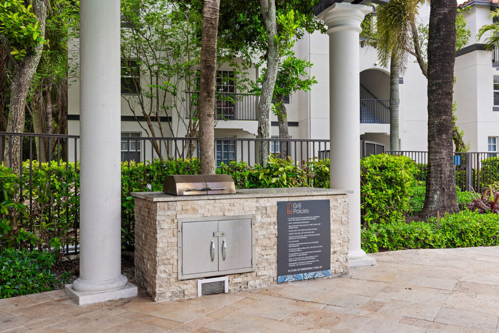 Outdoor stone grill station with a stainless steel grill, storage cabinet, and a sign displaying grill policies, surrounded by greenery, trees, and white pillars in front of an apartment building.