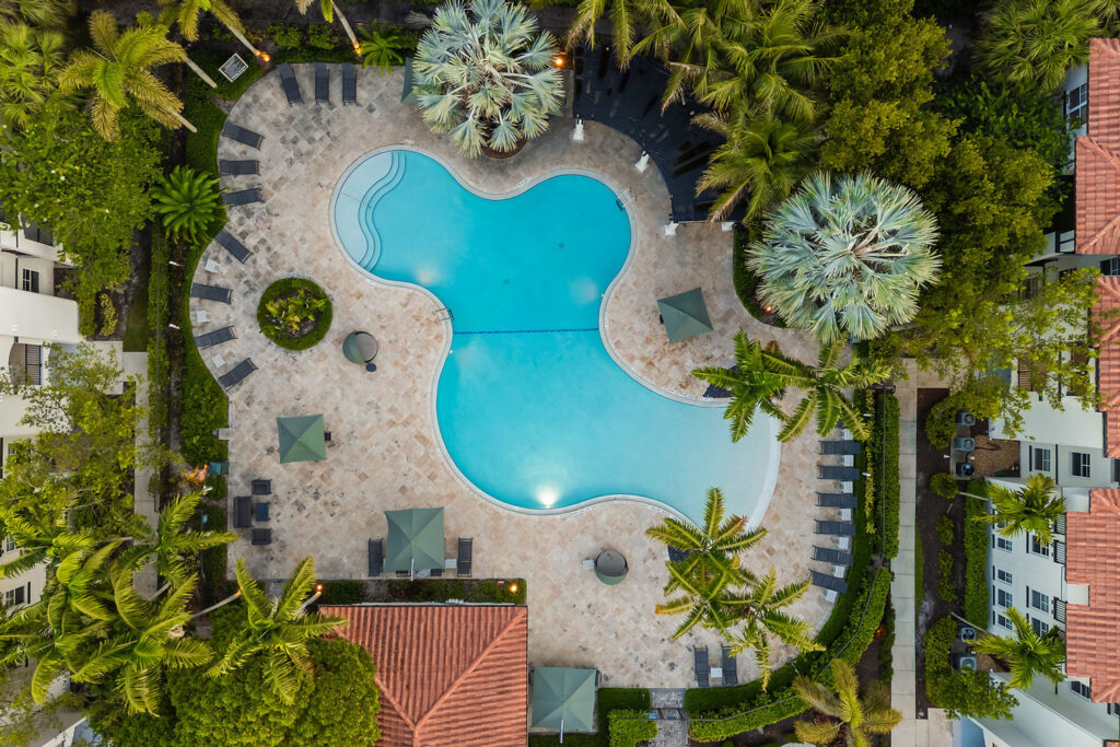 Aerial view of a kidney-shaped swimming pool surrounded by lounge chairs, tables with umbrellas, palm trees, and red-roofed buildings in a lush, tropical setting.