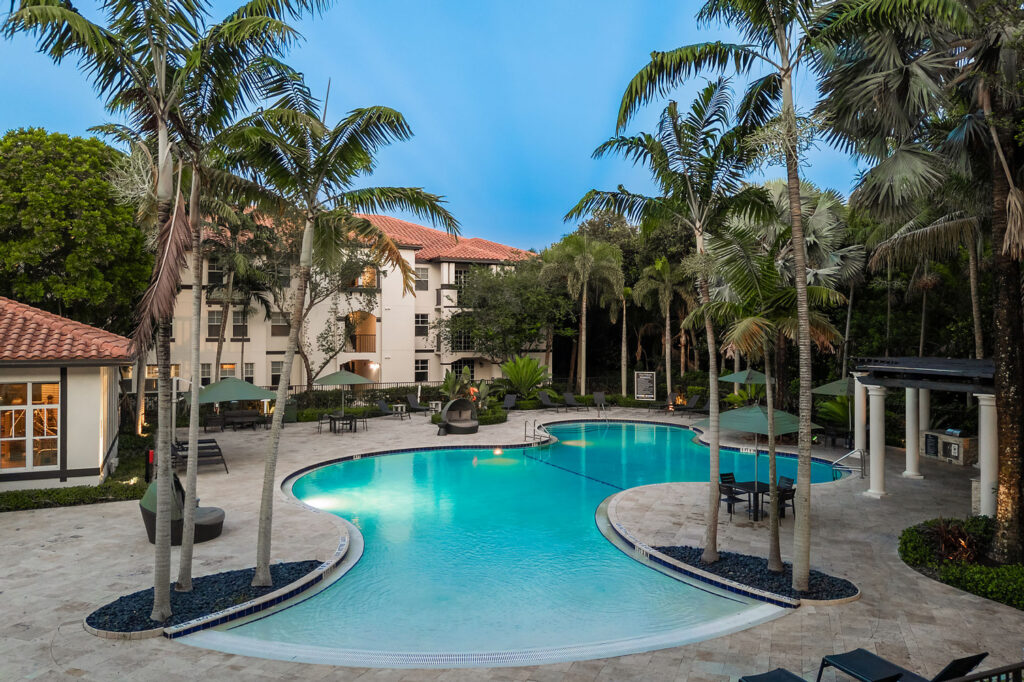 A resort-style outdoor pool with curved edges, surrounded by palm trees, lounge chairs, umbrellas, and a hot tub. Modern apartment buildings with red-tiled roofs are in the background under a clear blue sky.