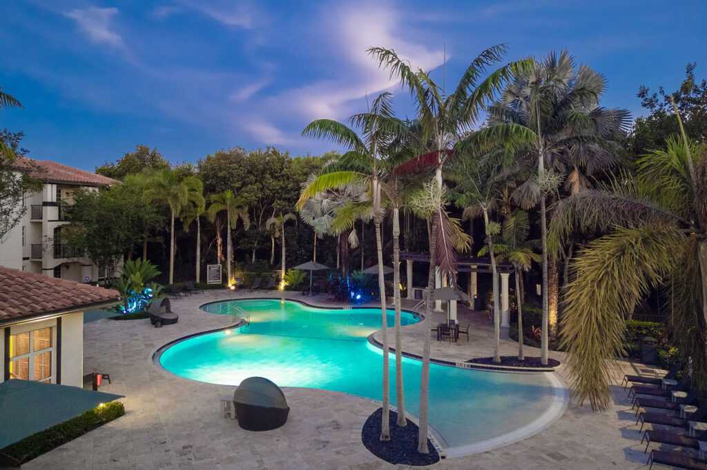 A resort-style swimming pool at dusk, surrounded by tall palm trees, lounge chairs, and tropical landscaping, with glowing blue water and soft outdoor lighting creating a serene, inviting atmosphere.