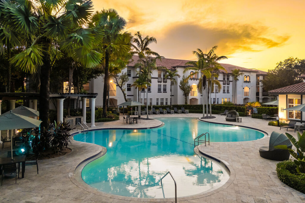 A resort-style swimming pool with palm trees, lounge chairs, and umbrellas, set against white apartment buildings at sunset, creating a relaxed, inviting atmosphere.