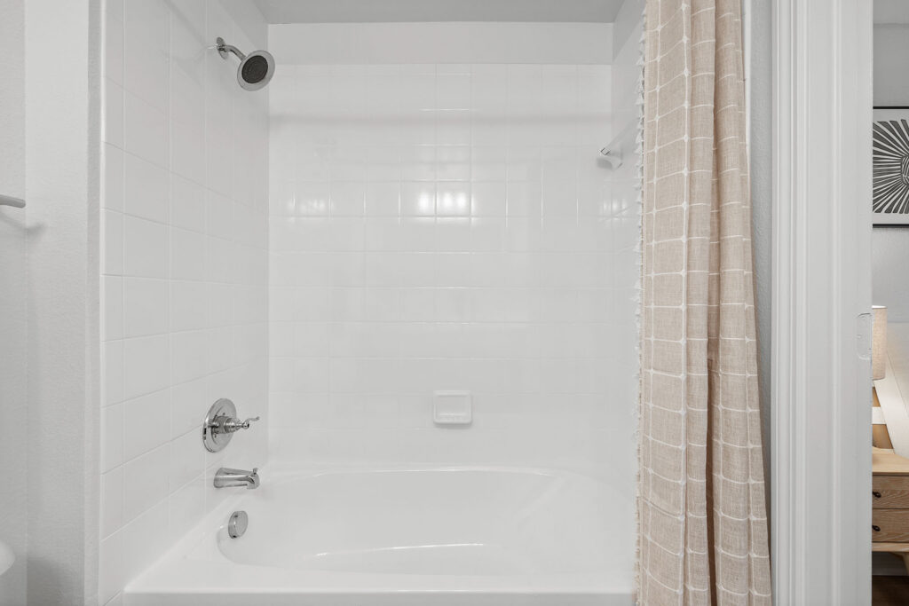 A clean, white bathtub with a tiled wall surround, silver faucet, and showerhead. A beige and white checkered shower curtain is partially drawn to the right. A small soap holder is built into the wall.