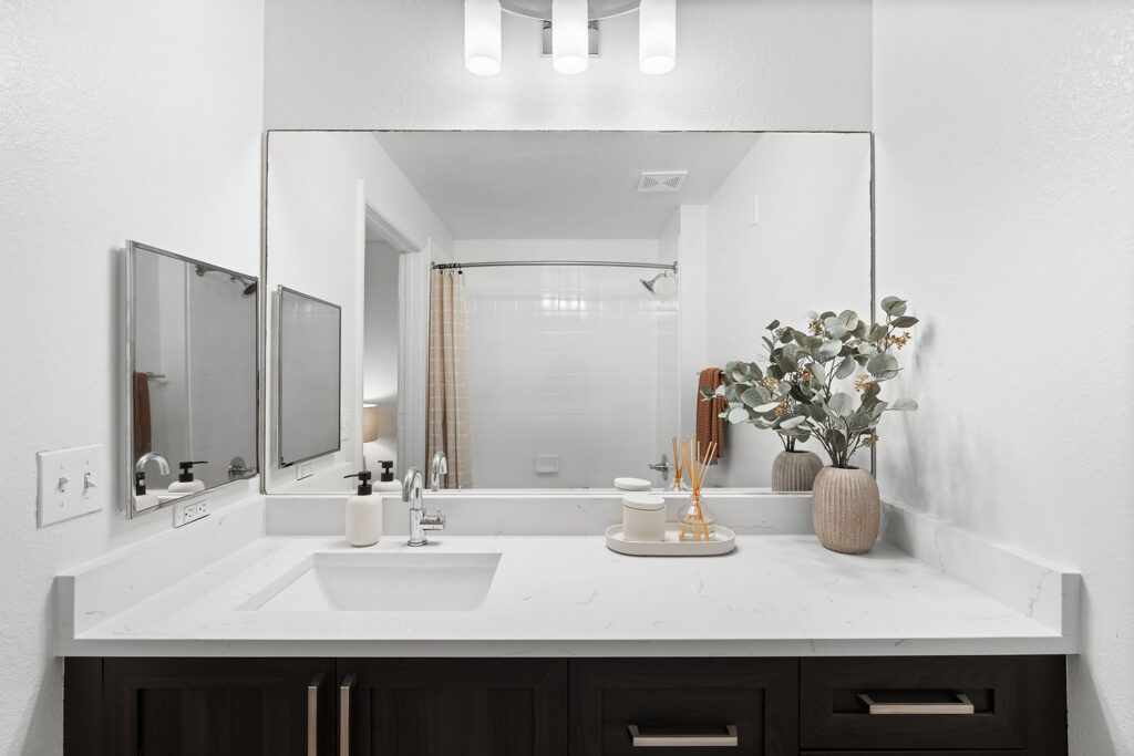 Modern bathroom vanity with white countertop, rectangular sink, large mirror, and dark wooden cabinets. Decor includes a vase with greenery, reed diffuser, soap dispenser, and towels. Shower with curtain in the background.
