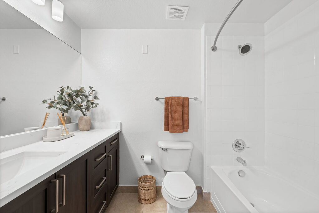 Modern bathroom with a white countertop, dark cabinets, and a large mirror. There is a toilet, a tub with a shower, a brown towel on a rack, and a potted plant on the counter for decoration.