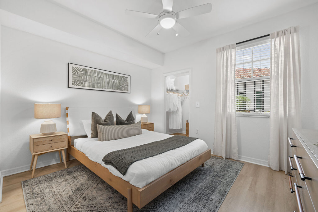 A modern bedroom with a wooden bed, white bedding, gray throw blanket, two nightstands with lamps, a dresser, wall art, and a window with sheer curtains letting in daylight. An open closet door is visible.