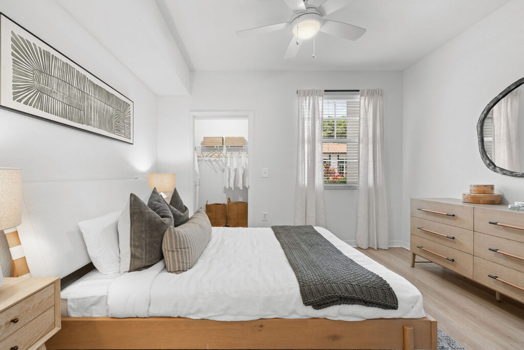A modern, bright bedroom with a wooden bed, white bedding, gray pillows, a gray throw, two side tables with lamps, a dresser, a wall mirror, a ceiling fan, and an open closet. Sunlight enters through a window with white curtains.
