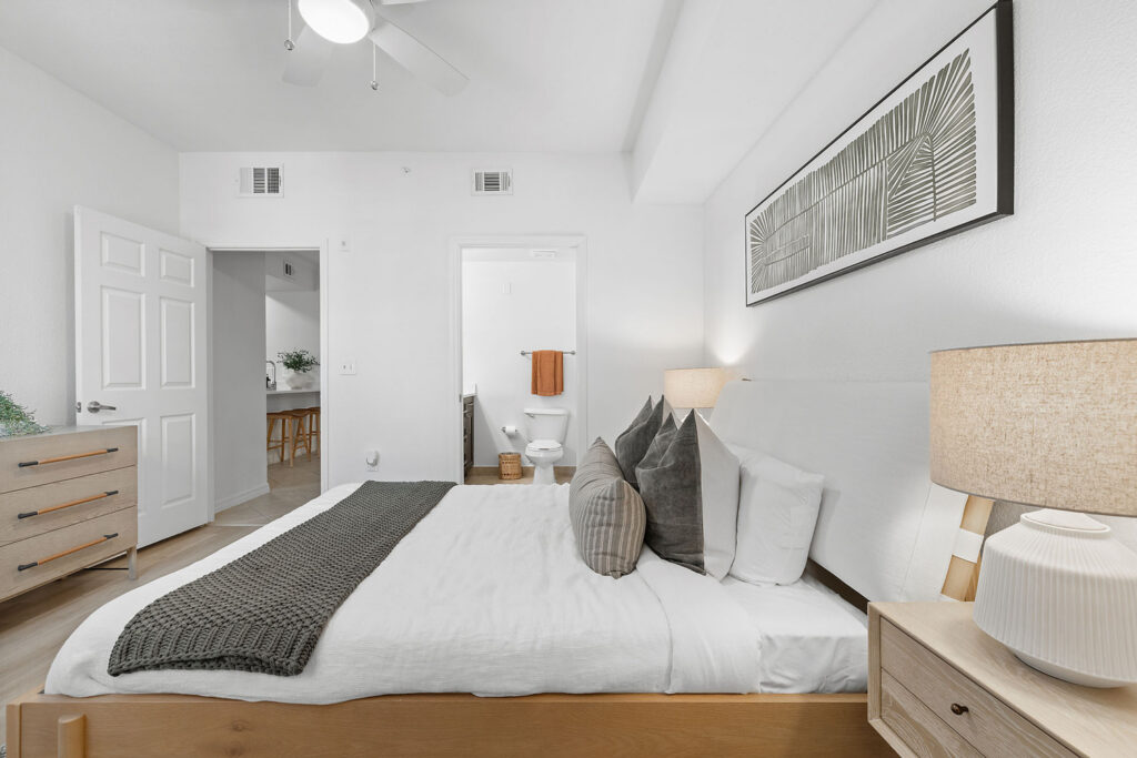 A modern bedroom with a wooden bed, white bedding, gray throw blanket, and several pillows. A dresser, nightstand with lamp, and framed leaf artwork decorate the space. An open door reveals a bathroom in the background.
