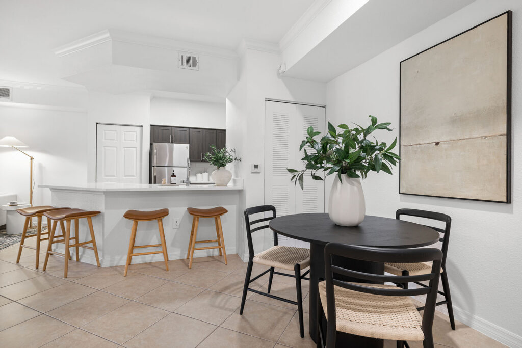 Modern kitchen and dining area with a round black table, four chairs, bar counter with four wooden stools, minimalist decor, large white vase with greenery, neutral wall art, tile flooring, and white walls.