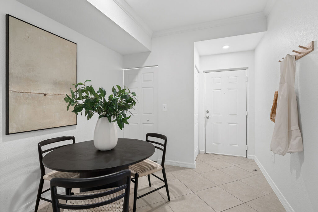 A minimalist entryway and dining area with a round black table, three chairs, a vase with green foliage, neutral wall art, tiled floor, a white door, and a wall hook holding a tote and a white cloth.