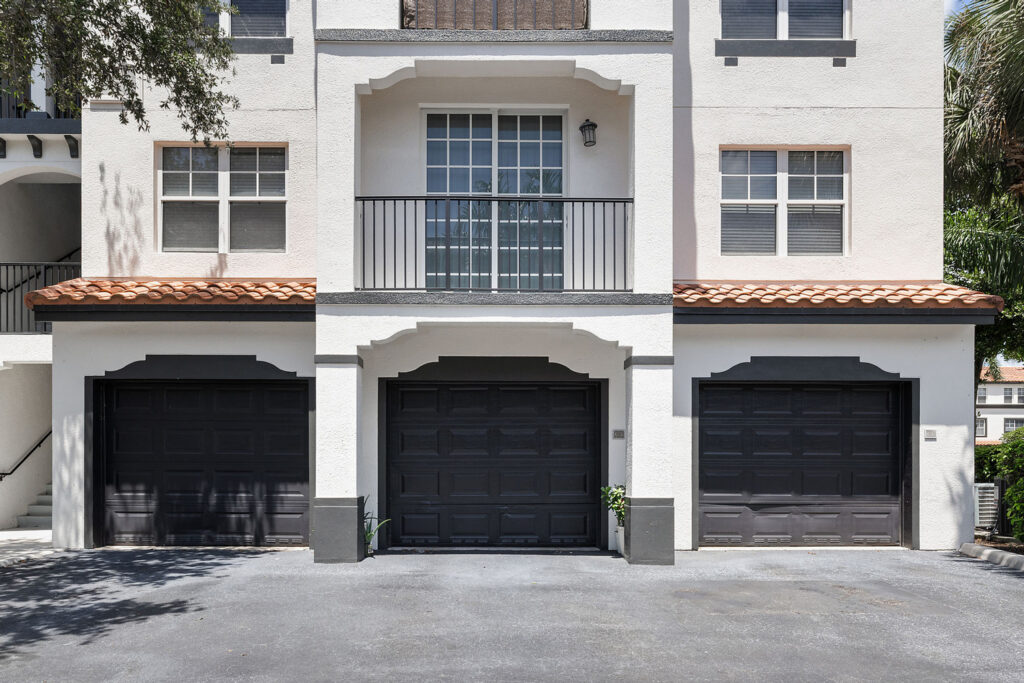 Three side-by-side black garage doors in a white, two-story residential building with a balcony and windows above the center garage. Green plants sit in front of the middle garage, and a tree branch is visible on the left.