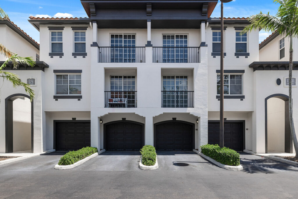 A modern three-story townhouse building with white walls, black trim, three black garage doors, small balconies on the second floor, and palm trees framing the entrance.