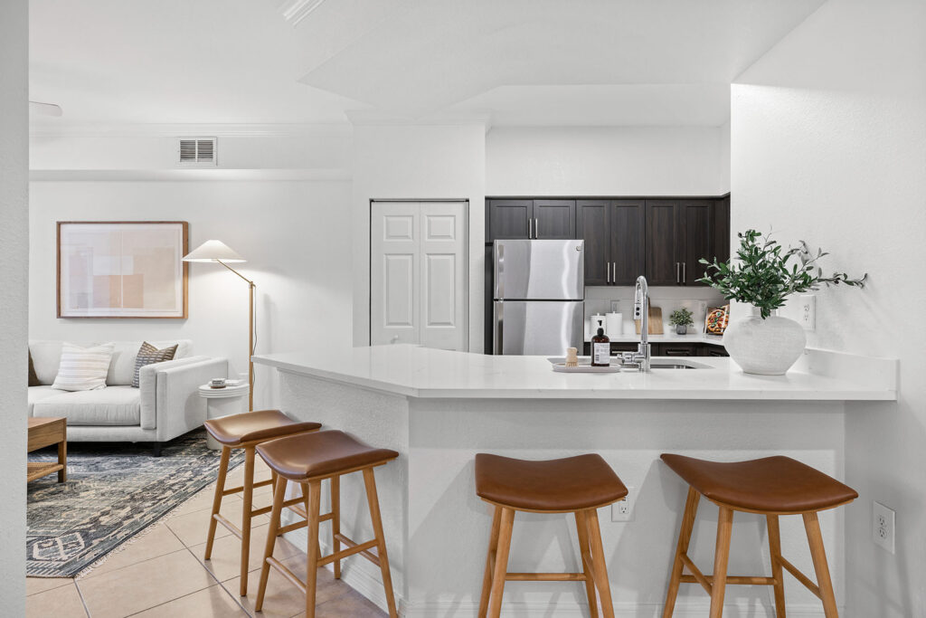 Modern kitchen with dark cabinets, stainless steel appliances, a white countertop with three brown barstools, and a living area in the background featuring a couch, framed art, and a floor lamp.