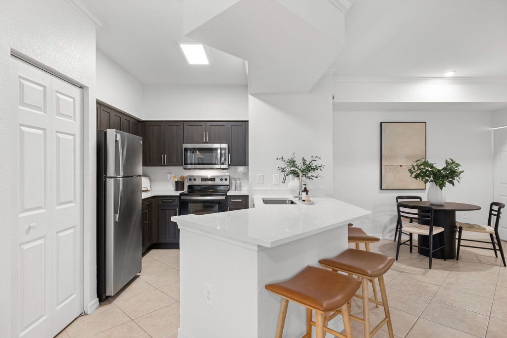 Modern kitchen with dark cabinets, stainless steel appliances, and white countertops; a breakfast bar with two wooden stools; adjacent to a small dining area with a round table and four chairs.
