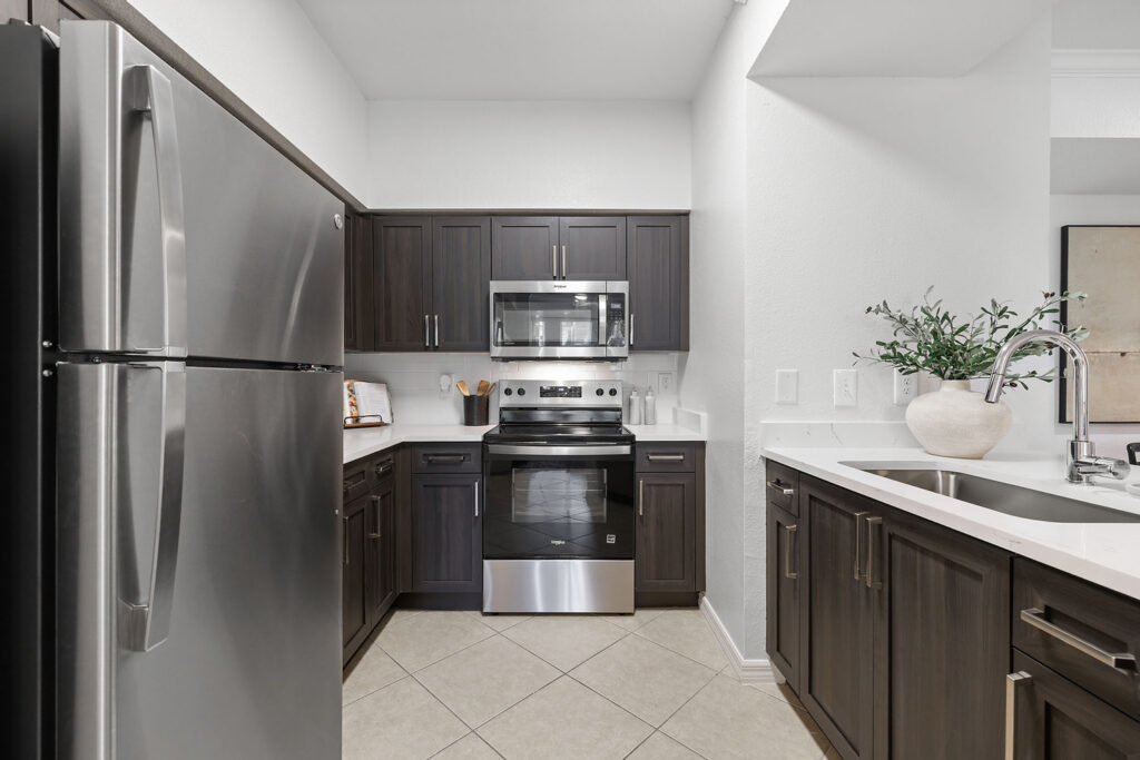 Modern kitchen with dark wood cabinets, stainless steel appliances, a refrigerator on the left, an oven with a microwave above it, a white countertop, a sink on the right, and a vase with greenery.