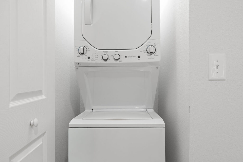 A white, stacked washer and dryer unit is placed in a small laundry closet with light-colored walls and a white door to the left. A light switch is visible on the right wall.