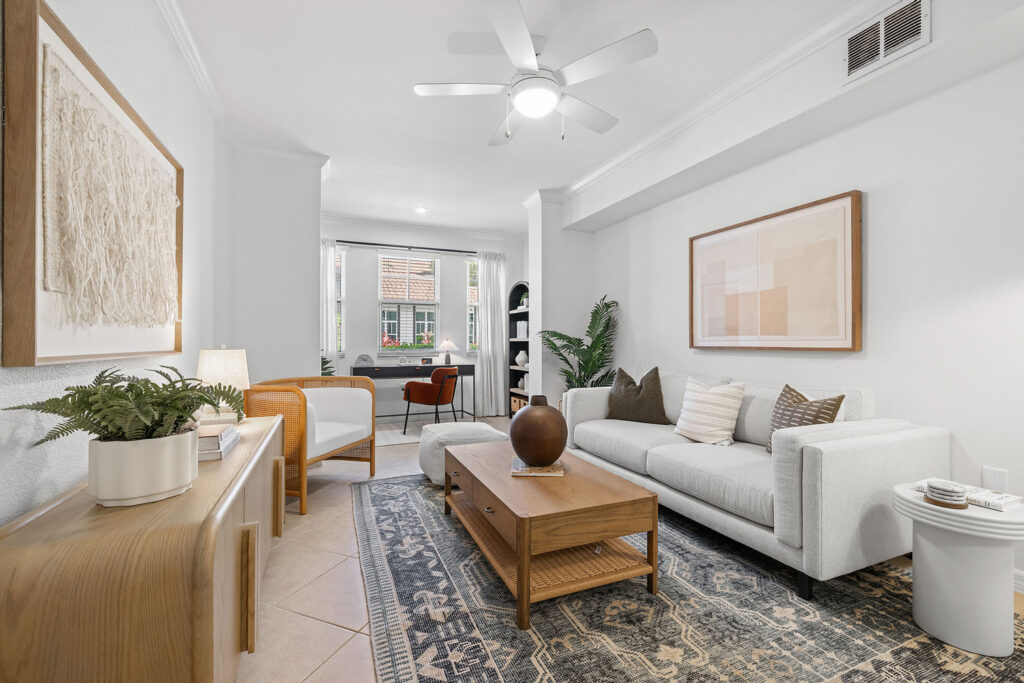 A modern living room with a white sofa, wooden coffee table, armchair, indoor plants, and framed art. A rug sits under the furniture and a ceiling fan is overhead. In the background, a desk faces large windows.