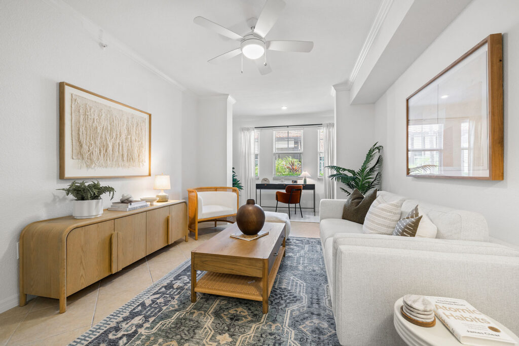 Bright living room with white walls, light furnishings, a white sofa, wooden coffee table, sideboard, framed artwork, indoor plants, and a ceiling fan. In the background is a window and a small workspace with a desk and chair.