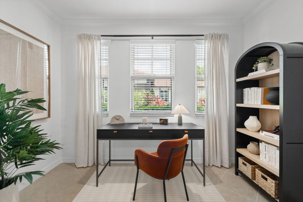 A modern, bright home office with a black desk, brown chair, lamp, decorative items, bookcase with books, and large windows with white curtains letting in natural light. A green plant sits in the left corner.