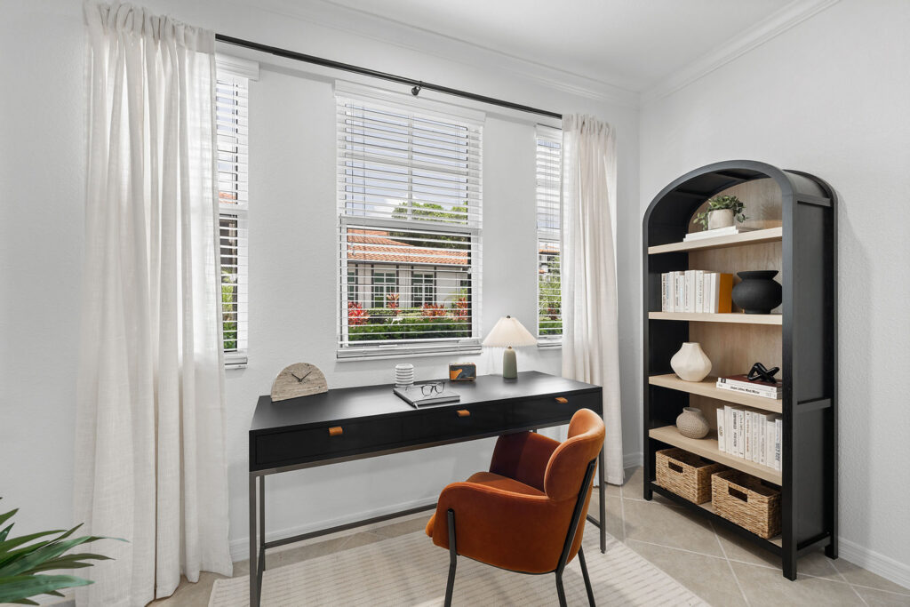 A modern home office with a black desk, orange chair, lamp, and decor. Large windows with white curtains let in natural light. A black bookshelf holds books, baskets, and decorative objects.