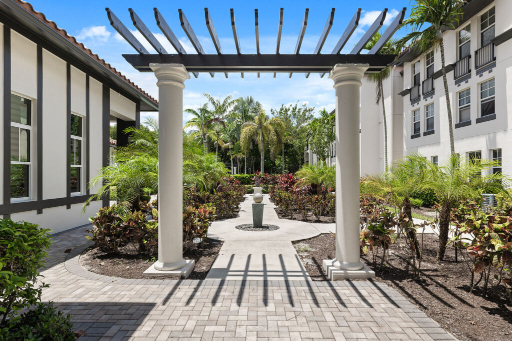 A courtyard with a paved walkway, tall white columns supporting a pergola, lush green palms, and landscaped plants between two white buildings under a bright blue sky.