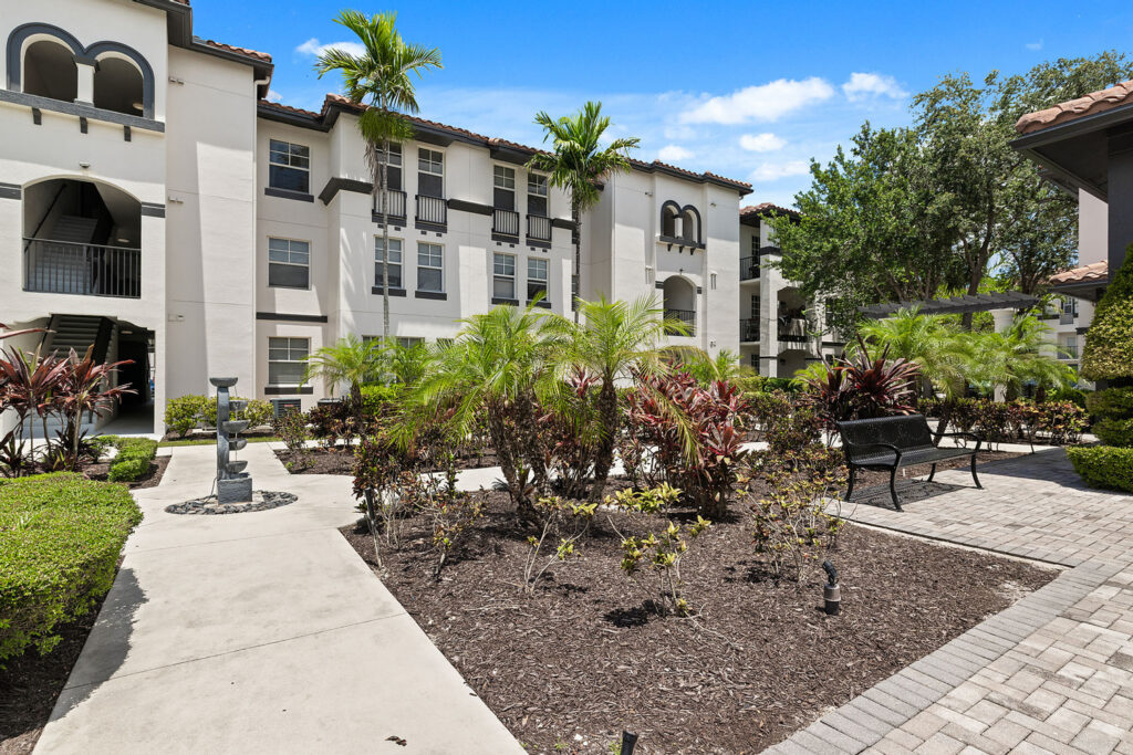 A sunny courtyard at an apartment complex with tropical plants, palm trees, a bench, a birdbath, and paved walkways surrounded by white, three-story buildings.