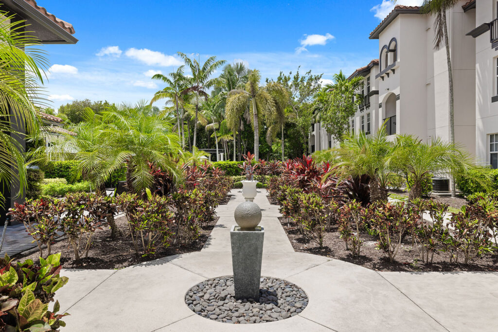 A landscaped courtyard with tropical plants, palm trees, and a decorative stone fountain centered on a paved walkway between two beige buildings under a bright blue sky.