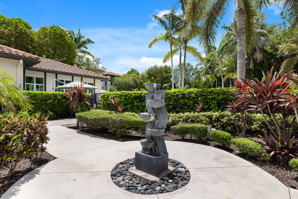 A modern tiered stone fountain stands surrounded by pebbles on a paved walkway, with manicured bushes, tropical plants, and palm trees near a house under a blue sky.