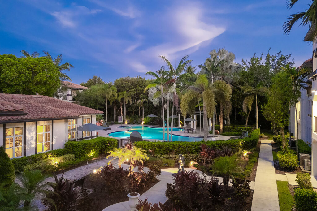 A landscaped courtyard with palm trees, gardens, and pathway leads to a pool surrounded by lounge chairs and umbrellas in the center of a residential complex at dusk.